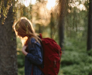 En flicka som går i skogen med en stor röd ryggsäck och solen lysande bakom träden