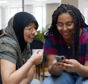 Två kvinnor sitter bredvid varandra, ler och tittar in i mobiltelefon.