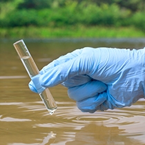 En hand med gummihandskar håller i ett provrör ovanför en vattensamling.