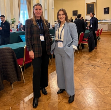 Två deltagare, kvinnor i kostym, står framför ett bord med flera deltagare