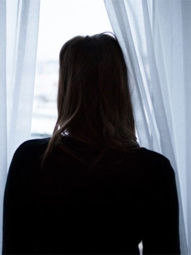 Rygg och bakhuvud på en kvinna som står och tittar ut genom ett fönster mellan ljusblå gardiner