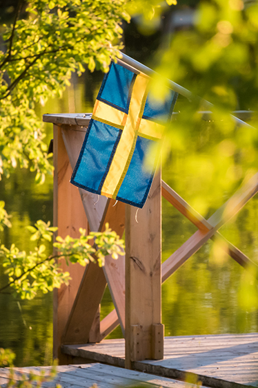 En svensk flagga hänger på ett trädäck vid vatten och grönska