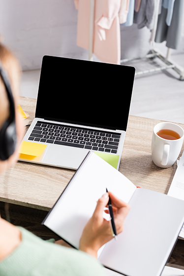 Bild över axeln på en person som sitter med hörlurar och skrivbok framför en laptop och en kopp te