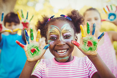 Ett leende barn med händer och ansikte målade i glada färger visar upp handflatorna, i bakgrunden står två barn till på samma sätt
