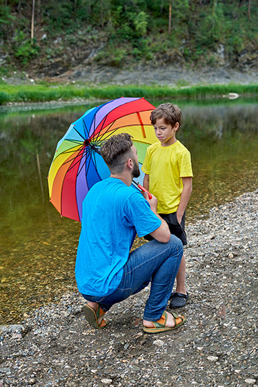 En pappa och en son pratar vid en damm under ett regnbågsfärgat paraply