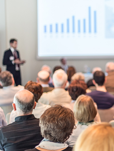 I bakgrunden syns en person i kostym hålla ett föredrag intill projektorbild med statistikstaplar, i förgrunden syns en sittande publik