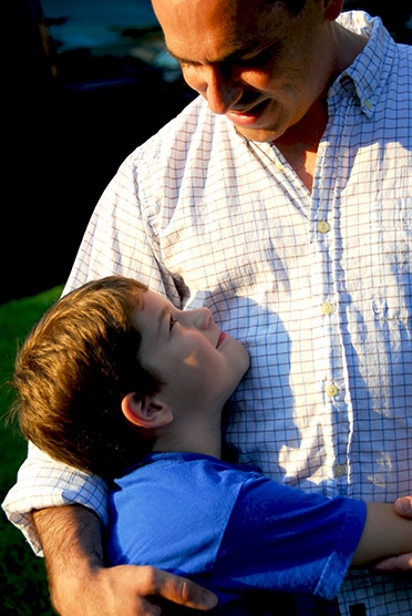 Ett barn kramar om sin pappa runt midjan och tittar upp på honom, båda ler mot varandra