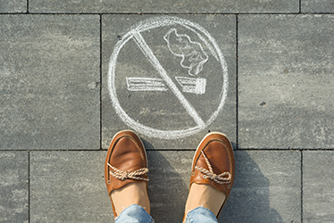 Ett par fötter mot marken, framför fötterna är en symbol ritad med vit krita, den föreställer en cigarett med ett streck över