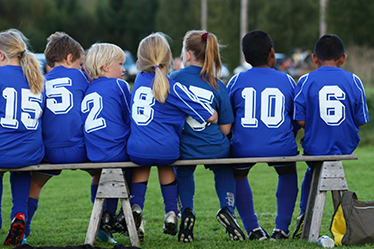 Sju barn i fotbollskläder sitter på en bänk vid sidan av gräsplanen, bilden är tagen bakifrån