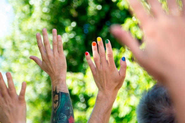 Händer som sträcks uppåt, några naglar är målade i regnbågens färger, i bakgrunden syns grönska