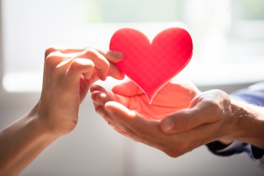 En hand ger ett rött pappershjärta till någon med kupade händer.