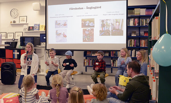 Kvinna håller sagostund för en grupp barn i bibliotek, bildskärm i bakgrunden.