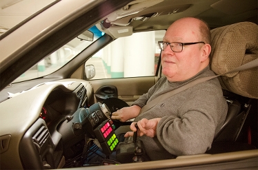 Man med glasögon och skalligt huvud sitter i förarsätet på sin anpassade bil. Han tittar på fotografen genom sidofönstret som är nedrullat