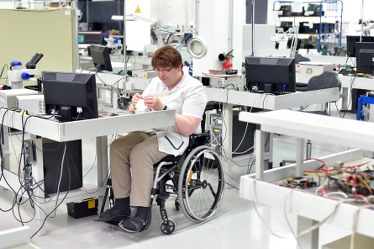 Ung man, användare av rullstol, sitter vid en arbetsbänk och gör något slags monteringsarbete. En dataskärm står på hans bord. Flera arbetsbänkar finns i rummet, men utan några som sitter vid dem. 