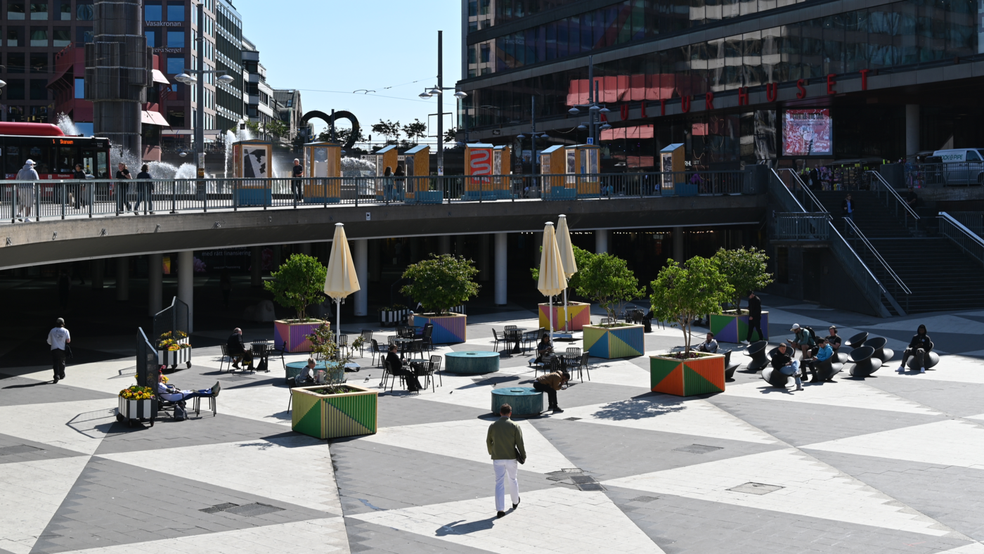 Livligt torg med människor, sittplatser och modern arkitektur i stadsmiljö.
