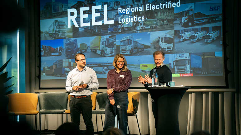 Tre personer deltar i en paneldebatt framför en skärm med texten "REEL Regional Electrified Logistics".