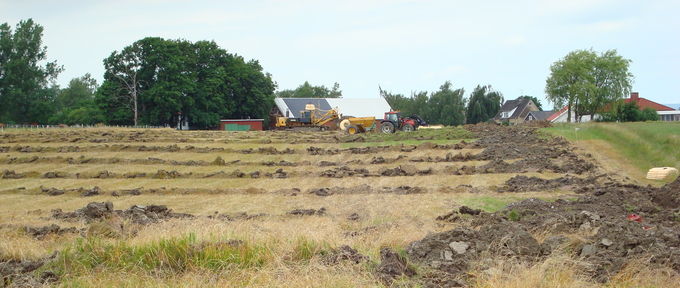 Åker med nygrävda diken. Traktor hus och träd i bakgrunden.