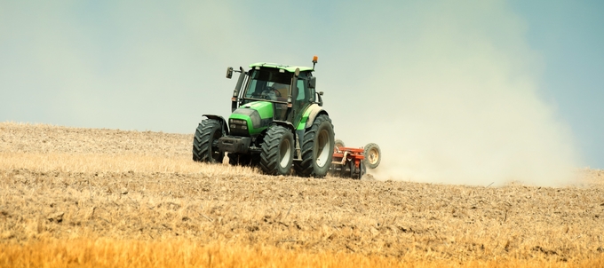 Grön traktor harvar ett fält under en klar himmel.