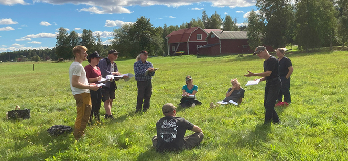 Grupp människor diskuterar utomhus på en äng med röd lada i bakgrunden.
