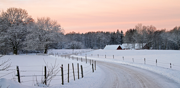 Vinterlandskap med snötäckt väg och röd lada vid solnedgång.