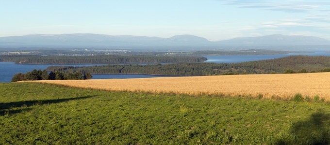Vy över gröna och gyllene fält mot en sjö och fjärran blå berg under klar himmel.