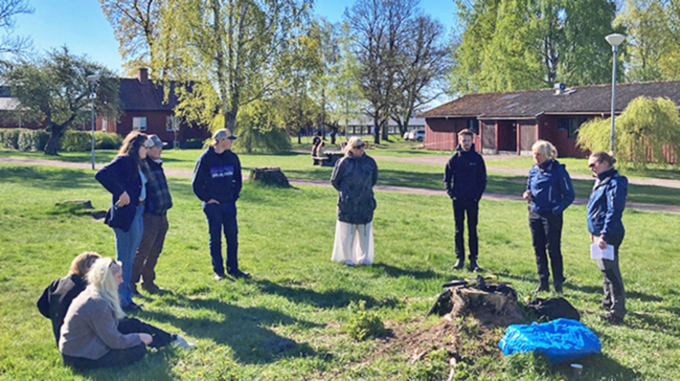 Flera personer står i en halvcirkel och pratar med träd och byggnader i bakgrunden