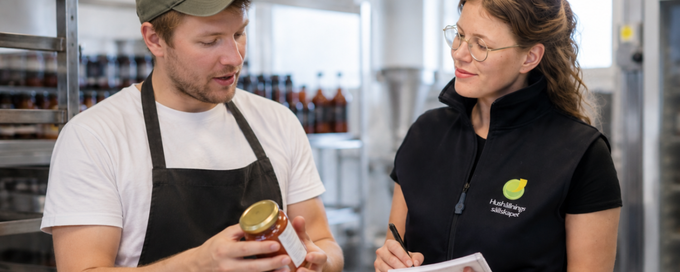 Två personer i en produktionslokal; en man i förkläde visar en burk medan en kvinna antecknar.