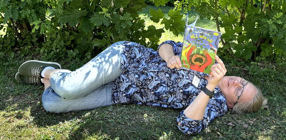 Eva från Infoteket har jeans och blå brokig blus. Hon ligger och läser en bok under ett träd i solskenet. Boken heter Autism inifrån, nej inte så!