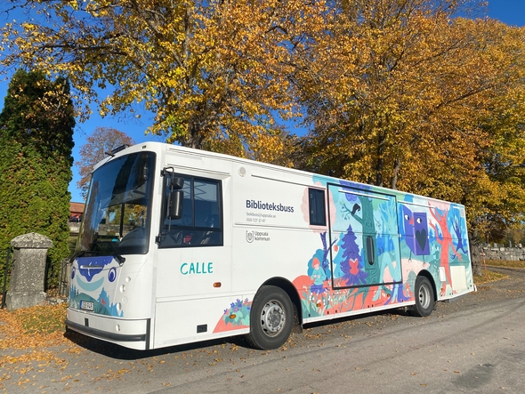 Vit biblioteksbuss med illustrationer i olika färger och texten Calle, med träd i höstfärger i bakgrunden.