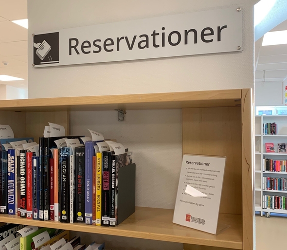 En bokhylla med böcker på rad som har vita lappar istoppade. Ovanför en skylt med texten Reservationer, och en bildsymbol bredvid med en hand som lyfter upp en bok.