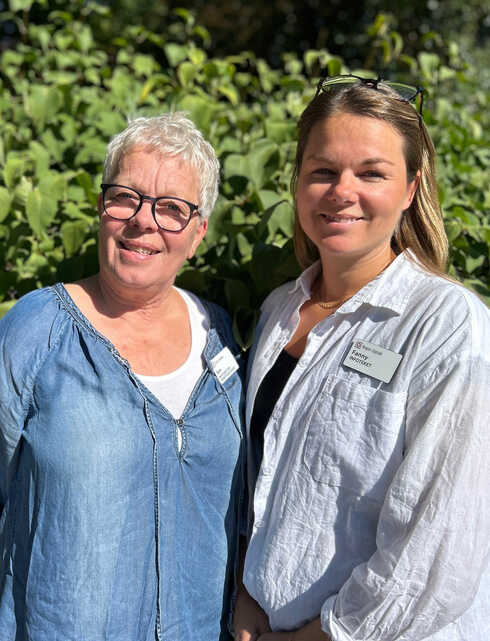 Ann och Fanny ler mot kameran i soligt väder framför en bakgrund av gröna blad.
