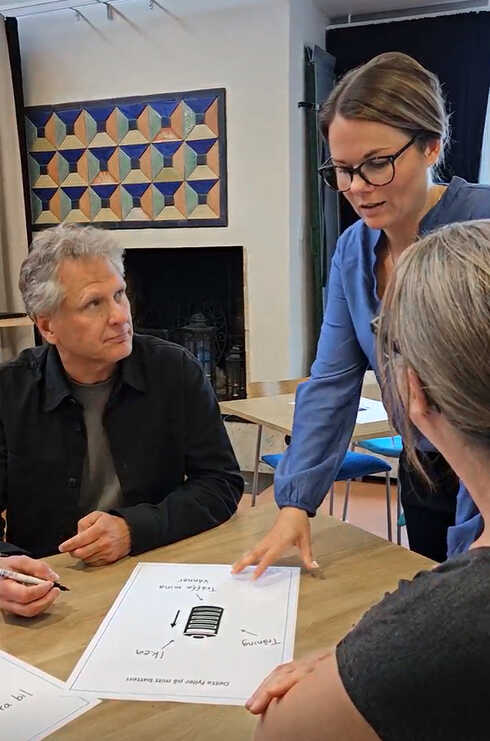 Person med glasögon presenterar ett grupparbete vid ett mötesbord för några andra personer.