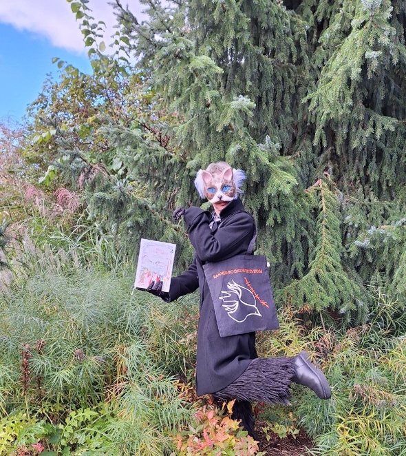 En figur med kattmask för ansiktet, fuskpäls och ytterrock står ute i en skog med en bok i handen och en svart tygkasse på axeln.