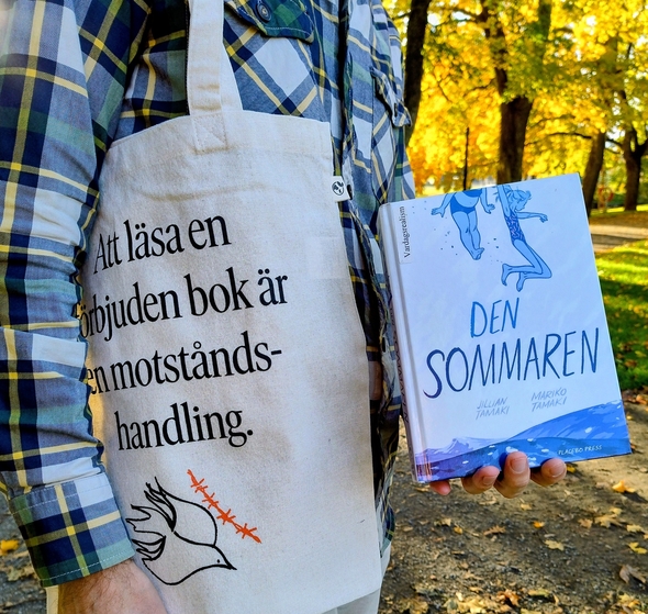 En person håller upp en bok med titeln Den sommaren. Personen har en naturvit tygkasse över axeln, med texten Att läsa en förbjuden bok är en motståndshandling.