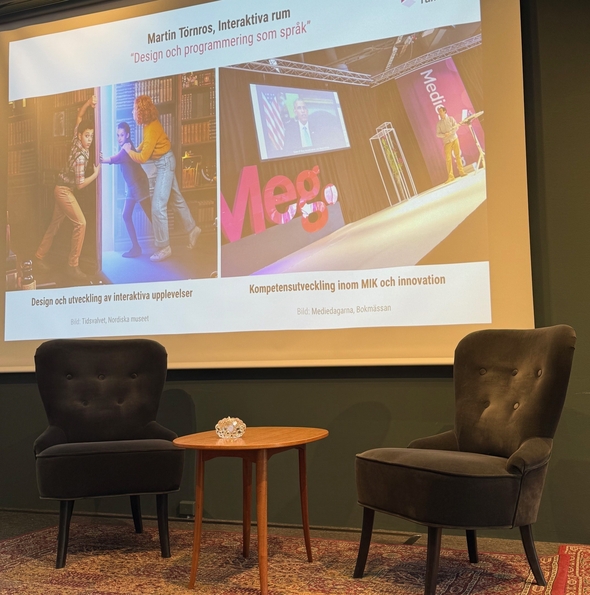 En projektorduk med två fåtöljer och ett bord på en scen nedanför. På duken visas en sida med texten Design och programmering som språk samt Design och utveckling av interaktiva upplevelser och kompetensutveckling inom MIK och innovation. 