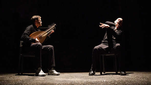 Två män på en scen där den ena spelar luta och den andra gestikulerar under en föreställning.