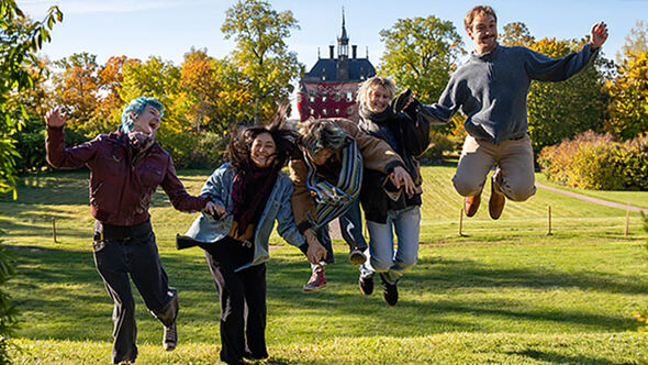Fem unga personer hoppar i gräset i en park med höstfärgade träd och ett slott i bakgrunden.