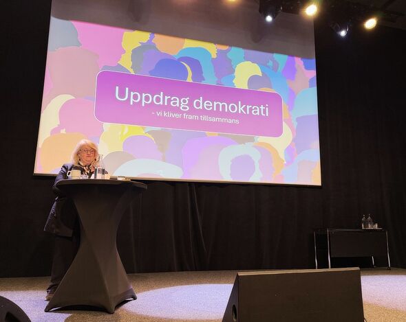 På en scen står en kvinna i ljust hår och glasögon bakom ett ståbord. I bakgrunden syns en projektorduk med texten Uppdrag demokrati - vi kliver fram tillsammans.