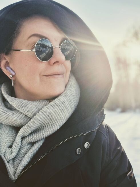 Närbild på leende kvinna i vinterjacka och halsduk med solglasögon, hörlurar och snöig bakgrund.
