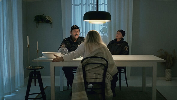 Två poliser sitter vid ett bord mittemot en person insvept i en filt under en taklampa.