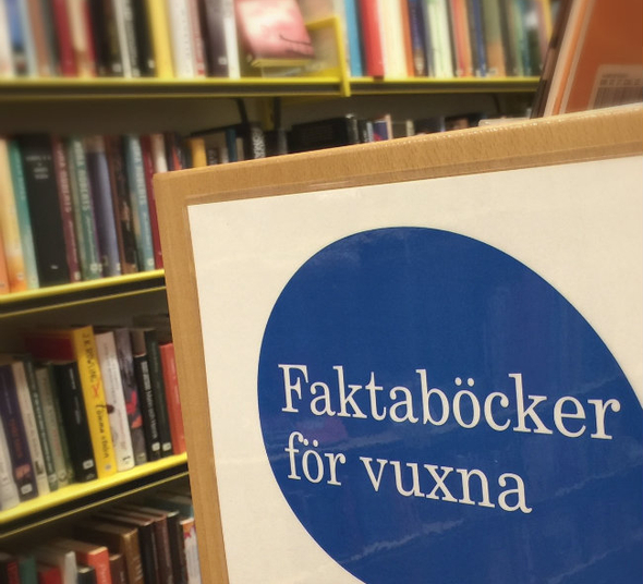 Skylt med texten faktaböcker för vuxna med bokhylla fylld med böcker i bakgrunden.