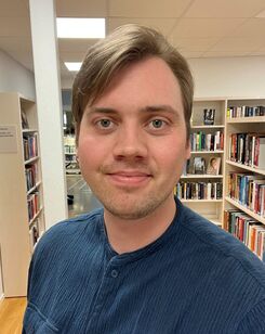 Porträttbild på Erik, som har brunt hår och blå skjorta. I bakgrunden finns flera bokhyllor.