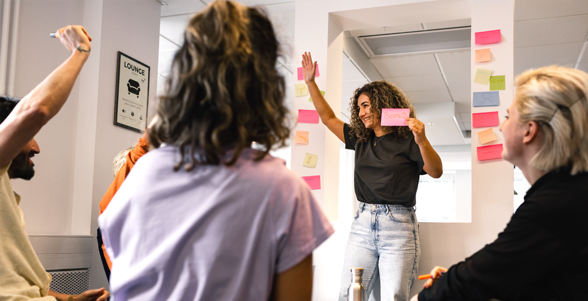 Grupp i workshop där en kvinna håller upp en post-it medan deltagare räcker upp handen