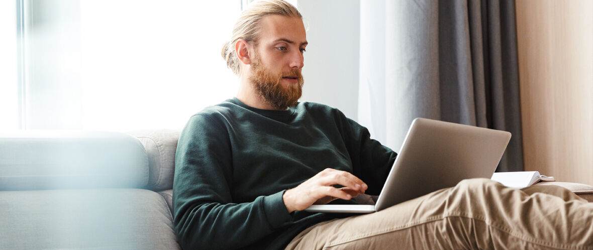 Person med skägg sitter i soffan och använder en laptop.