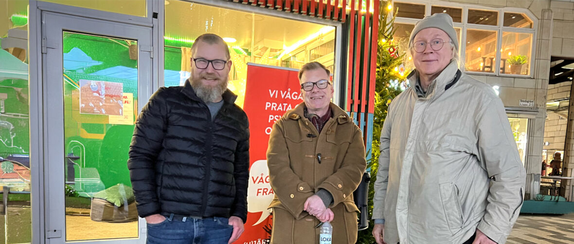 Tre män står leende utanför entré vid juldekorationer