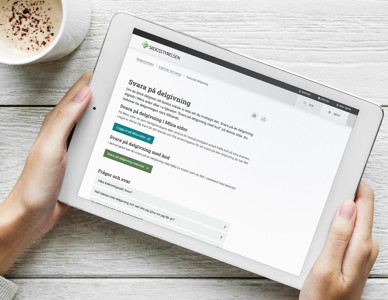 Två händer håller en surfplatta som visar ett webbaserat formulär, bredvid en kopp kaffe.