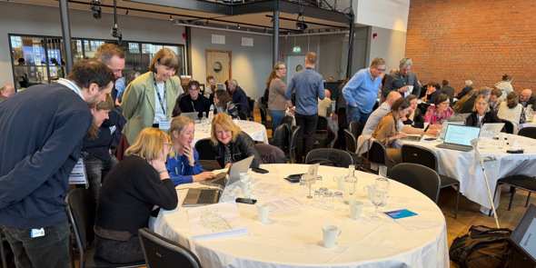 Deltagare i konferensen sitter vid runda bord och diskuterar med bärbara datorer.