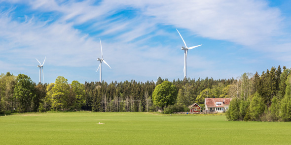 Grön åker med ett hus vid skog och tre vindkraftverk under blå himmel